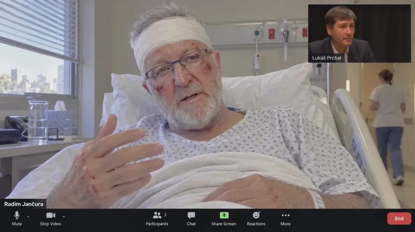 Záběr z videohovoru přes Zoom — Radim Jančura s obvázanou hlavou, hematomem na tváři a rukou v dlaze leží v nemocniční posteli. V pravém horním rohu — redaktor Seznam Zprávy Lukáš Prchal.